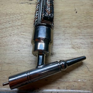 May include: A handcrafted metal beer tap with a decorative braided design in silver and copper tones. The tap features a polished chrome finish and a spout with a small pin at the end. The tap is designed for dispensing beer.