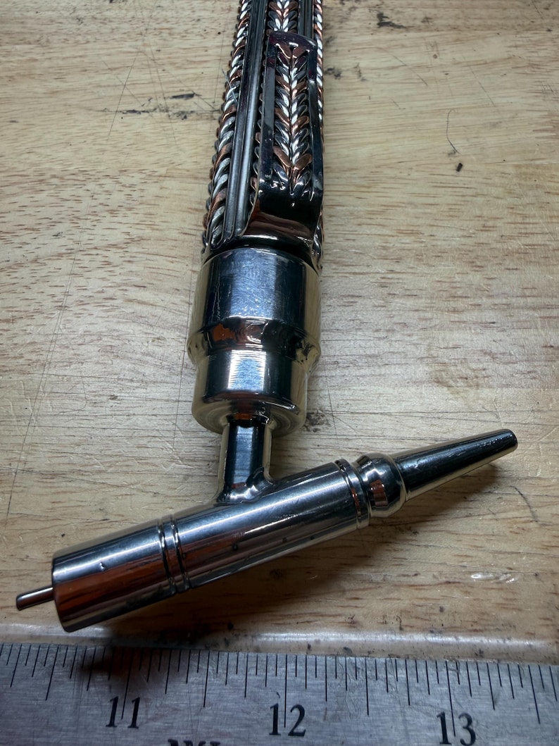 May include: A handcrafted metal beer tap with a decorative braided design in silver and copper tones. The tap features a polished chrome finish and a spout with a small pin at the end. The tap is designed for dispensing beer.
