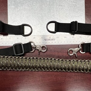 Puede incluir: Una eslinga de rifle de paracord negra y verde con dos correas ajustables negras y dos mosquetones plateados. La eslinga está colocada sobre una superficie de madera con una regla para la escala.