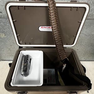 Pode incluir: Uma geleira Engel marrom aberta com interior branco e uma bandeja branca. Uma al&ccedil;a de ombro preta e uma al&ccedil;a tran&ccedil;ada marrom s&atilde;o vis&iacute;veis. A geleira cont&eacute;m uma al&ccedil;a preta e documentos. O logotipo Engel &eacute; vis&iacute;vel na tampa.