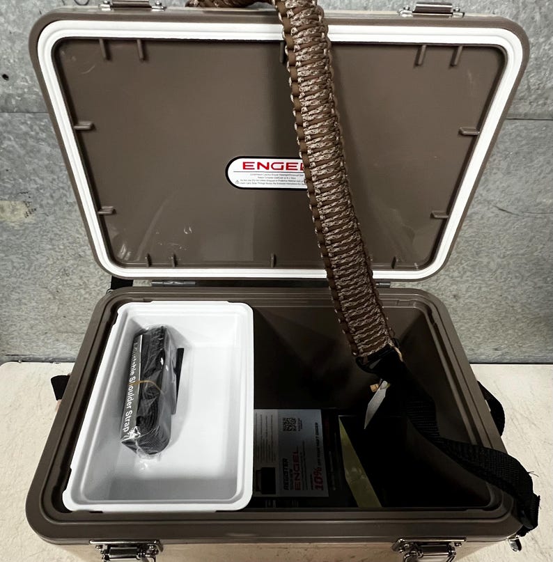 Pode incluir: Uma geleira Engel marrom aberta com interior branco e uma bandeja branca. Uma al&ccedil;a de ombro preta e uma al&ccedil;a tran&ccedil;ada marrom s&atilde;o vis&iacute;veis. A geleira cont&eacute;m uma al&ccedil;a preta e documentos. O logotipo Engel &eacute; vis&iacute;vel na tampa.