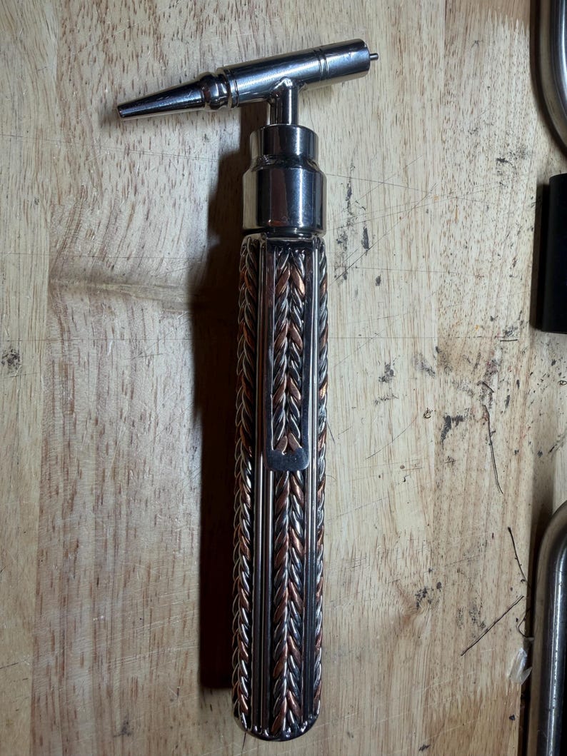 May include: A metal wine bottle opener with a braided copper and silver handle. The opener has a T-shaped design with a pointed tip. The handle is approximately 5 inches long.