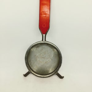 May include: A vintage tea strainer with a red plastic handle and a circular, fine-mesh stainless steel strainer. The handle has a hole for hanging. The strainer has small feet for stability.