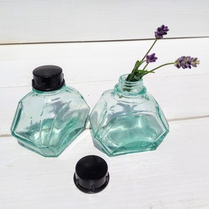 May include: Two clear glass ink bottles with black lids. The bottles have a faceted design and are sitting on a white surface. One bottle has a sprig of lavender flowers inside.