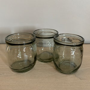 May include: Three clear glass candle holders with a dark grey rim. The candle holders are round and have a textured surface.