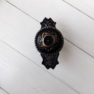 May include: Black and copper cabinet knob with a decorative design. The knob has a round shape and a raised center.