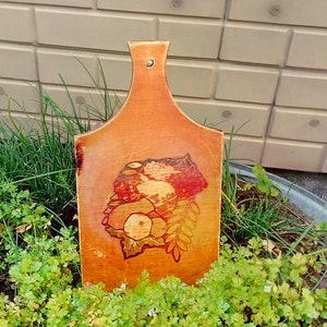 Può includere: Un tagliere di legno con un disegno dipinto a mano di frutta e foglie. Il tagliere è di colore marrone chiaro con una tonalità leggermente arancione. Il disegno è in tonalità di rosso, arancione e giallo.