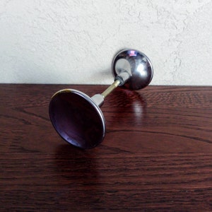 Może przedstawiać: Chromowa klamka do drzwi z okrągłą, płaską podstawą i okrągłym, lekko wypukłym wierzchołkiem. Klamka znajduje się na ciemnobrązowej drewnianej powierzchni.