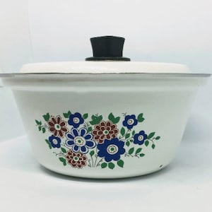 Könnte beinhalten: Weiße emaillierte Auflaufform mit einem schwarzen Knopf auf dem Deckel und einem Blumendesign in den Farben Blau, Grün, Braun und Rot.