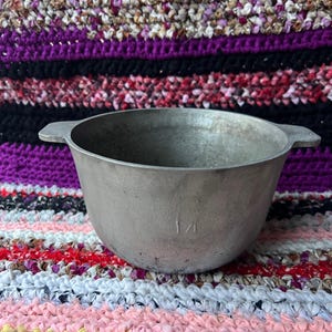 Puede incluir: Un pequeño cuenco de metal plateado con dos asas. El cuenco tiene forma redondeada y el número "1/4" está estampado en el lateral. El cuenco está sobre una tela a rayas coloridas.