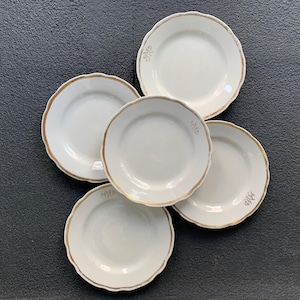 May include: Five white ceramic plates with scalloped edges and gold trim. Each plate has a gold monogram. The plates are arranged on a dark surface. A set of vintage tableware.