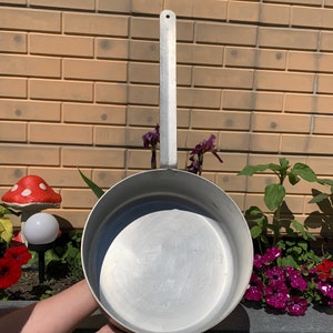 May include: A large, silver, aluminum cooking pot with a long handle. The pot is empty and has a smooth, shiny surface.