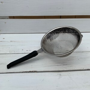 Puede incluir: Un colador de malla plateado con un mango de plástico negro. El colador está sobre una superficie de madera blanca.