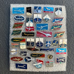 Pode incluir: Uma coleção de alfinetes de avião vintage em várias formas e cores. Os alfinetes apresentam desenhos de aviões e texto cirílico, incluindo "CCCP" e números de modelos de aviões como "TU-134" e "IL-62".
