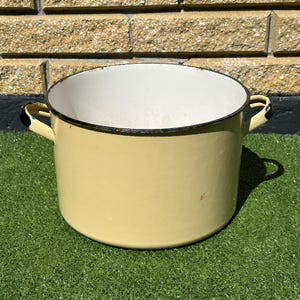 May include: A vintage yellow enamelware pot with a white interior and black trim. The pot has two side handles and sits on a green surface. This cooking pot is ideal for kitchen decor or use.