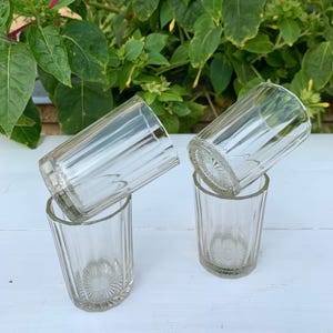 Puede incluir: Cuatro vasos transparentes de estilo vintage. Cada vaso tiene estrías verticales y un motivo floral decorativo en la base. Los vasos están dispuestos en pares, con dos vasos apilados sobre los otros dos.