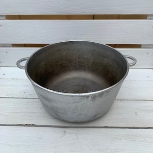 May include: A large, silver aluminum pot with two handles. The pot is empty and has a worn, aged appearance.