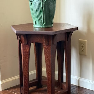 May include: A green ceramic vase with a textured design sits atop a dark brown, hexagonal wooden side table. The table has four curved legs and a lower shelf. The vase has two handles and a detailed floral pattern.