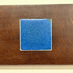 May include: A rectangular wooden plaque with a dark brown finish. The plaque features a recessed square with a textured blue surface, framed by a white border. The wood grain is visible.