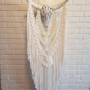 May include: White macrame wall hanging with a white, carved, bull skull in the center. The macrame features a fringe design with tassels.