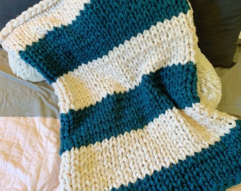 May include: A chunky knit throw blanket with white and teal stripes. The blanket is folded and draped over a bed.