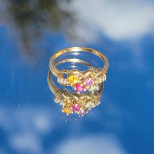Puede incluir: Un anillo de oro con un grupo de piedras preciosas de colores, incluyendo piedras amarillas, rosas y azules, y pequeñas piedras transparentes. El anillo se refleja en una superficie espejada.