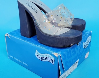 May include: A pair of navy blue platform sandals with clear straps adorned with iridescent rhinestones. The chunky heels and platform soles are navy blue. The sandals are displayed on a blue Skechers box.
