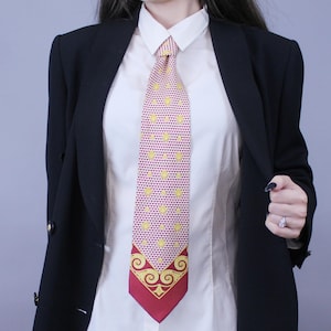 May include: A patterned tie with a burgundy and yellow design. The tie features a repeating pattern of yellow hearts on a burgundy background. The bottom of the tie has a burgundy border with a gold ornate design. The tie is worn with a white collared shirt and a black blazer.