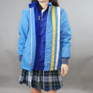 May include: A light blue jacket with a white and yellow stripe design on the front. The jacket has a blue fleece collar and a zipper closure. The person is wearing a blue polo shirt and a green and blue plaid skirt.