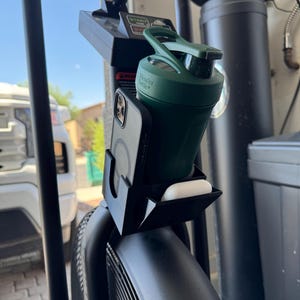 May include: A black exercise bike with a phone holder, a green BlenderBottle, and a pair of white earbuds. The phone holder is attached to the bike's console. The BlenderBottle is dark green and has a white logo.