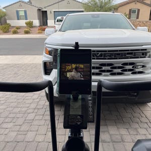 May include: A black exercise bike with a tablet holder attached to the handlebars. The tablet is displaying a video of a man in a green shirt with the text "The Joe Rogan Experience". The bike is parked in a driveway with a white pickup truck in the background.