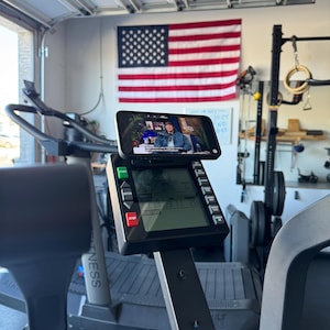 May include: A treadmill with a smartphone mounted on the console displaying a video. The console has a digital display showing workout metrics. An American flag hangs in the background.