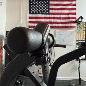 May include: Black exercise equipment with a curved handle and adjustable knob. An American flag hangs in the background. The equipment appears to be part of a home gym setup.