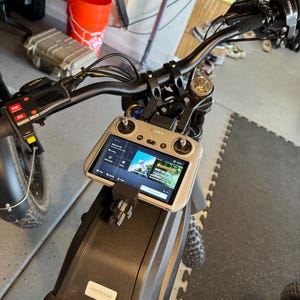 Puede incluir: Una motocicleta negra con un manillar negro y un mando de control de dron blanco y gris montado en el manillar. La pantalla del mando muestra la palabra "Running" y un mapa. La motocicleta tiene un asiento negro con una pegatina blanca que dice "PREORDER".