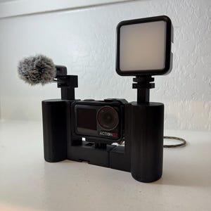 May include: A black video recording rig with a camera, microphone, and LED light. The camera is labelled "ACTION 5". The microphone has a fluffy windscreen. The LED light is square and emits a soft glow. The rig is designed for handheld use.