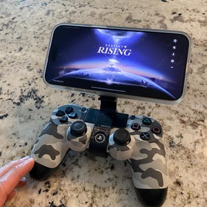 Puede incluir: Un mando de videojuegos con estampado de camuflaje con un smartphone adjunto, que muestra el juego "Destiny Rising". El mando tiene botones y joysticks negros. El teléfono se sujeta con un soporte negro.