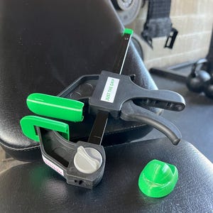 May include: A green and black clamp with a black handle and a gray knob. The clamp has a green label that says "JUST THE GRIP".