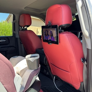 May include: Interior car shot with red leather seats and a tablet mounted on the headrest. The tablet displays a video. A child car seat and a small trash can are also visible.