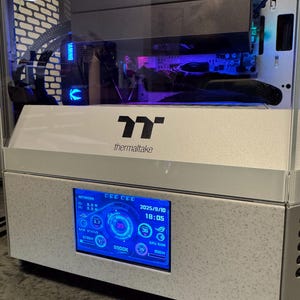 Może przedstawiać: Srebrna obudowa komputera Thermaltake z cyfrowym wyświetlaczem pokazującym informacje o systemie. Wyświetlacz pokazuje status sieci, użycie procesora i pamięci RAM oraz datę i godzinę. Obudowa ma przezroczysty panel boczny.