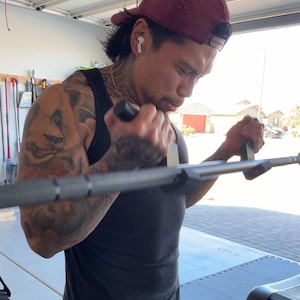 May include: A man wearing a maroon baseball cap and a black tank top is working out with a cable machine. He has tattoos on his arms and is using both hands to pull the cable down.