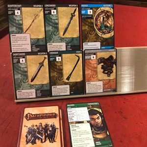 Op de afbeelding: Een Pathfinder Adventure Card Game-set met verschillende kaarten die op een rood fluwelen oppervlak zijn gelegd. De kaarten bevatten illustraties van wapens, bepantsering en andere voorwerpen. De kaarten hebben tekst die de naam van het voorwerp, de controle om te verkrijgen en de kracht van het voorwerp bevat.