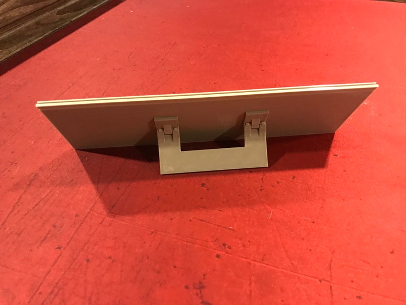 Op de afbeelding: Een beige plastic fotolijst met een standaard aan de achterkant. De lijst is ontworpen om rechtop te staan op een tafel of plank.