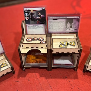 May include: A wooden organizer with four compartments for holding cards and tokens. The organizer has a Marvel logo on the front. The compartments are filled with cards and tokens, including a card with the text "Underground Distribution" and a card with the text "Alien Arms Factory".