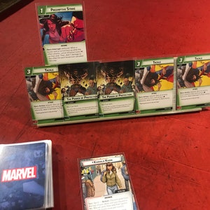 Op de afbeelding: Een verzameling Marvel-verzamelkaarten met personages zoals Kamala Khan en Wolverine. De kaarten worden getoond op een groene en witte standaard met de tekst "The Power of Protection" op twee van de kaarten.