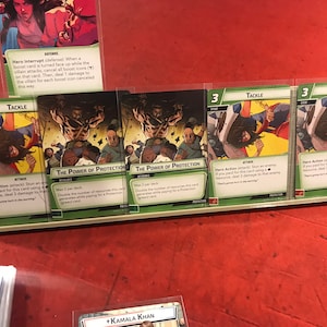 Op de afbeelding: Vier groene en witte kaarten voor een kaartspel met de tekst "Tackle" en "The Power of Protection". De kaarten bevatten illustraties van een vrouw in een superheldenpak.