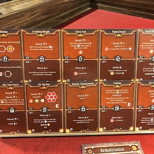 Op de afbeelding: Een set van tien bruine en oranje kaartspelkaarten met illustraties en tekst. De kaarten zijn gerangschikt in twee rijen van vijf. De kaarten hebben titels zoals "Windup", "Crushing Weight", "Extra Fuel", "Piston Punch", "Knock Out the Support", "One-Two Punch", "The Big One", "Robotic Enhancement", "Explode", en "Fire Bombs".