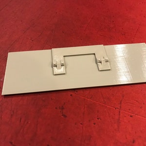 Op de afbeelding: Een rechthoekige witte plastic plaat met twee kleine grijze plastic scharnieren aan de bovenkant. De plaat ligt op een rood oppervlak.