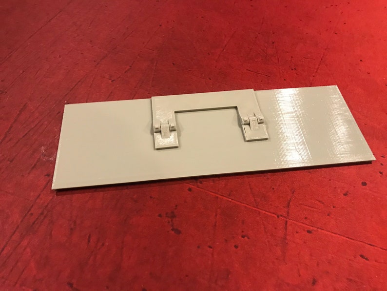 Op de afbeelding: Een rechthoekige witte plastic plaat met twee kleine grijze plastic scharnieren aan de bovenkant. De plaat ligt op een rood oppervlak.