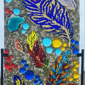 May include: A rectangular mosaic panel featuring a large blue and green peacock feather design alongside smaller yellow, red, and multicolored feathers. The artwork incorporates various glass beads, polished stones, and small square tiles.