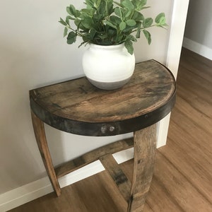 May include: A half-round wooden table with a metal band around the edge. The table is made from reclaimed wood and has a rustic look. A white ceramic planter with a green plant sits on the table.